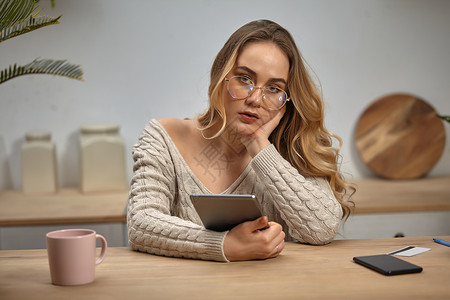 戴眼镜的博主女士 米色毛衣 拿着平板电脑 坐在厨房里的木桌旁 上面放着智能手机 塑料卡和粉色杯子 特写针织的高清图片素材