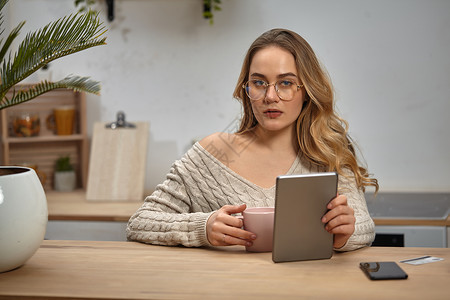 戴眼镜的女博主 米色毛衣 拿着平板电脑 粉红色的杯子 坐在厨房里的木桌旁 手掌 智能手机 塑料卡 特写药片高清图片素材