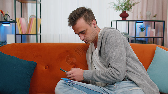 男人坐在沙发上使用信用卡和智能手机在网上购物时转账图片