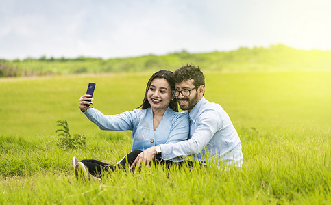 微笑的情侣坐在草地上自拍 年轻的情侣在田野里自拍 恋爱中的人用智能手机在田里自拍背景图片