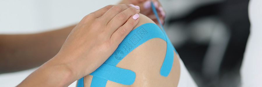 蓝色阔腿裤生理治疗师在门诊部的病人膝盖上应用动脉病录象带背景