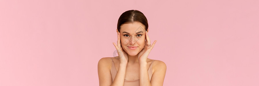 皮肤护理 美丽的脸触摸健康的面部皮肤的女人 微笑的女模特看着镜头 在粉红色的背景下微笑 网页横幅背景图片