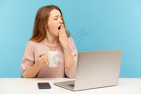 情感年轻女性在蓝背景的笔记本电脑上工作活力员工女士杯子办公室失眠时间咖啡唤醒就寝懒惰的高清图片素材