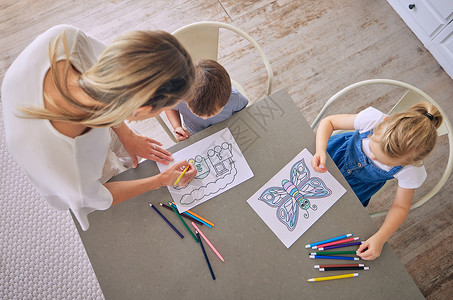 上图是小女孩和男孩坐在桌旁 拿着彩色铅笔和图片 妈妈在帮忙涂色 有两个孩子的白人母亲享受教育消遣和创造力背景图片