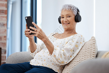 一个在沙发上用智能手机和耳机的 高龄女人 在家中的沙发上玩耍真实的高清图片素材