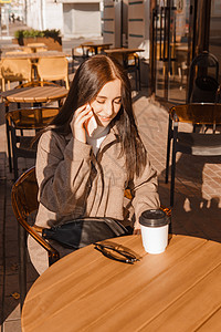 时髦的黑发女人坐在咖啡和手机室外咖啡厅的桌子上 女孩在电话里说话 秋天散步姿势青年情感微笑衣服热饮城市杯子摄影商业喝高清图片素材