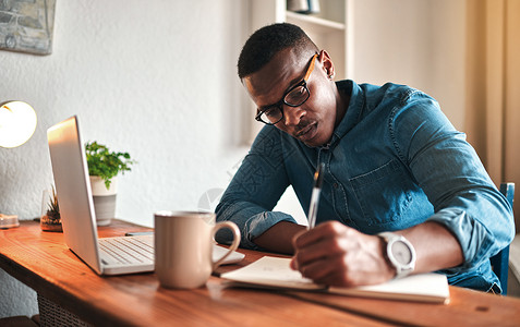 非洲裔美国企业家在笔记本上写作并规划项目战略 年轻人在他的创业公司远程工作 在家里用笔记本电脑工作 同时在书中安排约会背景图片