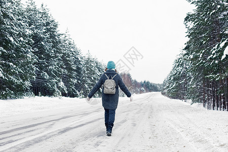 一个女孩穿着灰色大衣和蓝色编织帽子 背上背着灰色背包 走在冬季路边雪路高清图片素材