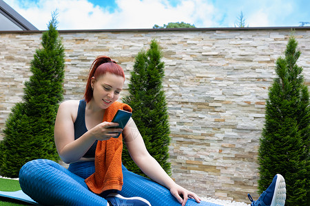 迷人的年轻健身女孩坐在户外的普拉提球上看着她的智能手机 女孩一边锻炼一边在她的社交网络上分享 健康和保健的概念青年说明运动装护理背景图片