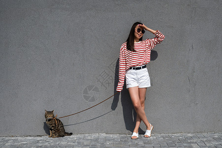 散步女孩和猫一位年轻女子带着一只灰色的小猫走在一条皮带上爪子哺乳动物朋友们城市叶子流浪汉公园尾巴猫科虎斑背景