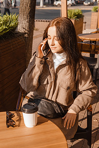 时髦的黑发女人坐在咖啡和手机室外咖啡厅的桌子上 女孩在电话里说话 秋天散步热饮杯子城市生活衣服长发摄影商业情感女士咖啡杯成功的高清图片素材