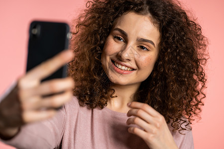 带着卷发在智能手机上做自拍的双胞胎微笑的快乐女孩 他们用粉红色的背景 技术 移动设备 社交网络的概念信息高清图片素材