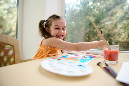 画画笔画快乐的学龄前小女孩 微笑地看着相机 同时用水彩画画画和油漆笔画画背景