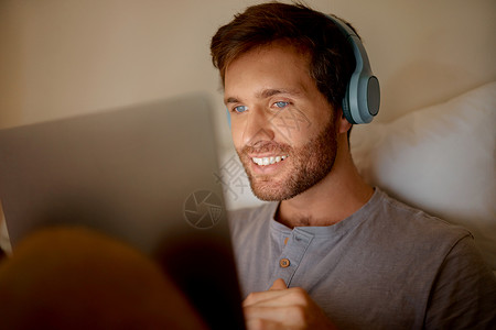 男人在数字平板电脑上在线播放电影 在卧室里看技术系列 晚上在家里的床上用耳机听音频 微笑着阅读社交媒体帖子的人串流高清图片素材