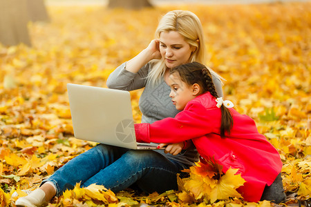 秋天简单女孩都市妇女和女儿在公园里带着笔记本电脑 穿着牛仔裤的苗条时髦女人使用笔记本 使用通信技术远程工作和环保生活方式的自由职业者落叶女性背景