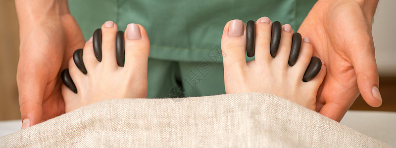 在温泉疗养所 女性按摩师手中的女脚趾之间用黑石头进行脚底按摩修脚岩石福利护理中心毛巾保健享受奢华皮肤背景