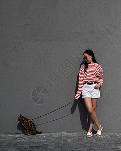 散步女孩和猫一位年轻女子带着一只灰色的小猫走在一条皮带上朋友们城市宠物猫咪虎斑冒险叶子马具女士毛皮背景