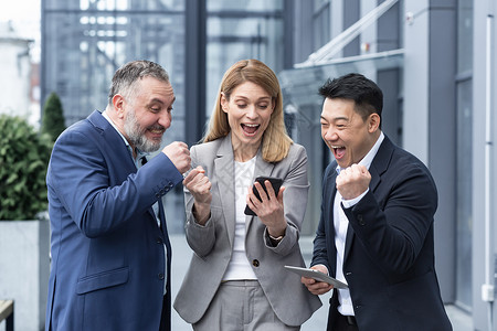 一群同事员工阅读智能手机的好消息 快乐的团队在使用 浏览手机时为成功的投资而高兴 站在办公楼附近聪明的高清图片素材