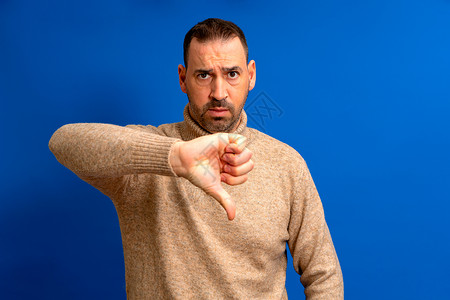戴棕色领头衣的有胡子的拉蒂诺男子看上去不开心 愤怒地表示拒绝和消极的拇指向下缩手势不喜欢高清图片素材
