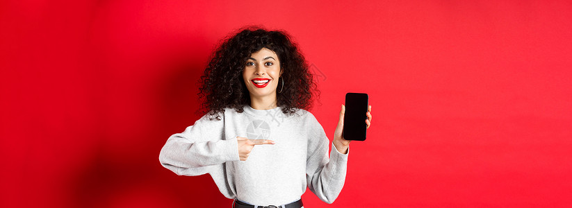 漂亮的女模特展示空的智能手机屏幕 指着手机显示屏微笑 站在红色背景下工作室女性促销技术广告购物电话女孩卷曲黑发吸引人的高清图片素材