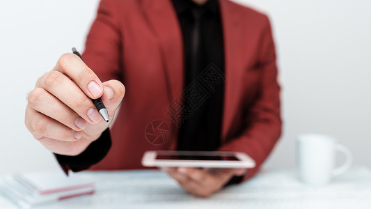 穿着红色外套的男子坐在白桌边 用笔指着重要信息 持有手机职业教育数位板金融创造力文档桌子企业家经理成人背景图片