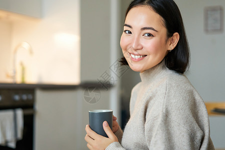 韩国饮料带着咖啡 喝热茶和在家舒适的笑笑的亚洲女孩的肖像 用饮料来暖和起来桌子沙发闲暇互联网女性太阳生活长椅自拍厨房背景