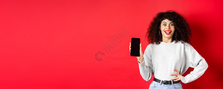 带着卷发和红嘴唇的兴奋女性 显示空智能手机屏幕 欢乐地尖叫 站在红色背景上女孩卷曲电话购物女性工作室生活广告黑发技术年轻的高清图片素材