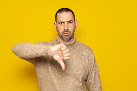 留着胡子的西班牙裔男子身穿米色高领毛衣 在黄色背景下表情严肃 不赞成地竖起大拇指象征高清图片素材