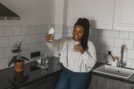 快乐的现代非洲裔美国年轻女孩谈论视频聊天并坐在家里交流的概念 微笑的时髦女孩在家里用电话制作自画像互联网相机房间女性手机卷发技术自拍高清图片素材
