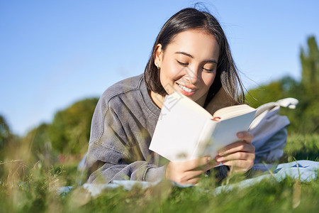 可爱的韩国女孩的画像 在公园里躺在草地上读书 手里拿着最喜欢的书放松 开心地笑着微笑成人花园女性背包快乐幸福黑发乐趣野餐亚洲高清图片素材