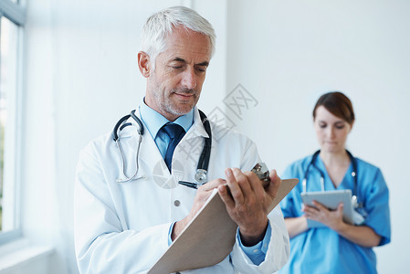 使用新旧方法检查记录 一位成熟的医生在他的剪贴板上检查记录 一名护士在后台浏览数字平板电脑背景图片