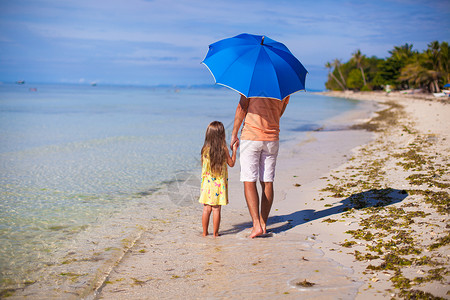 年轻父亲和他的小女儿在白沙滩上 在蓝伞下行走 轻松 太阳背景图片