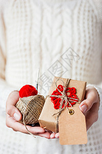 present穿着白色针织毛衣和手套的女人拿着一份礼物和一卷卷红线和亚麻绳 礼品采用手工钩编红色雪花牛皮纸包装 Present 的文本有一个空背景