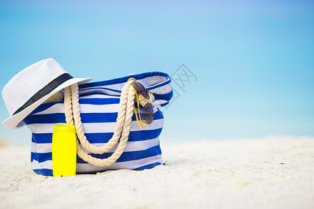 白沙滩上的蓝袋 草帽 墨镜和防晒霜瓶 海岸线背景图片