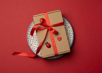 长方形棕色纸板盒 用丝绸红丝带绑在圆陶瓷红色板上 庆祝 假期背景图片