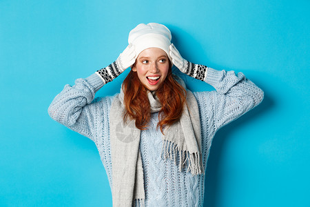 快乐的红发女孩戴着大米帽 围巾和手套 看着正对面微笑 站在蓝背景下站立 学生 舒适背景图片