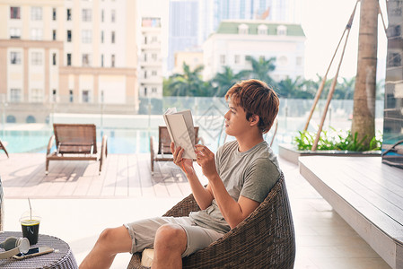 一个年轻的亚洲人正在户外泳池附近放松 手里拿着一本书 户外的 旅行幸福高清图片素材