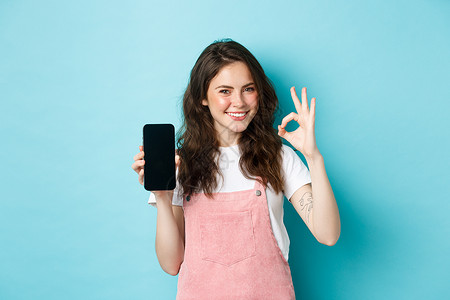 推荐网上商店或移动应用程序的现代时尚女孩的画像 在空的智能手机屏幕上显示好的标志 点头表示赞同 微笑满意 蓝色背景背景图片
