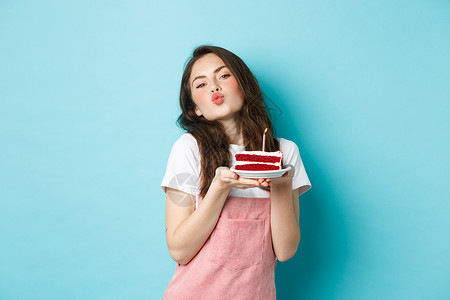 节日和庆祝活动 可爱的傻女孩在生日蛋糕上许愿 亲吻脸 庆祝生日 在蓝色背景下欢快地站着 学生 派对咧着嘴笑高清图片素材