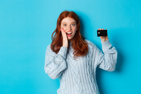 蓝色毛衣穿着毛衣的可爱红发女孩 展示信用卡 微笑着镜头 站在蓝背景上 工作室 新年背景