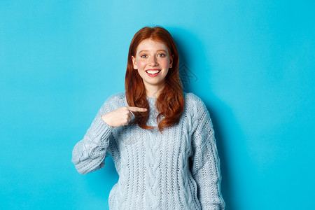 美丽的红发美女指着自己 笑着快乐 被选中 穿着蓝色的毛衣站在蓝背景上 舒适背景图片