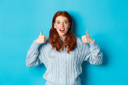 穿着毛衣的红发女孩惊艳 展示拇指举起和赞美产品 看着摄影机惊讶 站在蓝背景上站立 促销 快乐的背景图片