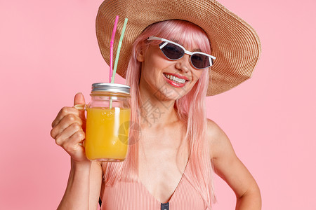戴着草帽 泳装和太阳镜的快乐女人的画像 对着镜头微笑 喝着热带果汁鸡尾酒 在粉红色的工作室背景中与世隔绝地摆姿势嘴高清图片素材