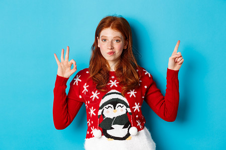 圣诞快乐 穿着Xmas毛衣的红头红发女孩 指向右上角 展示新年宣战和赞成 赞美产品 蓝色的 年轻的背景图片