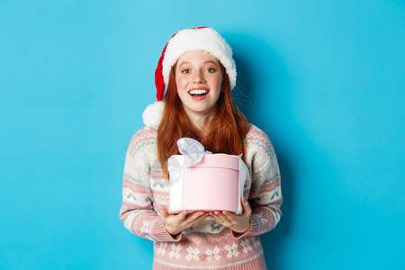 冬季和庆祝活动的概念 快乐的红发女孩收到圣诞礼物并表示感谢 拥抱着带礼物的盒子 微笑着 站在蓝色背景中的圣诞帽中 冬天 发型背景图片