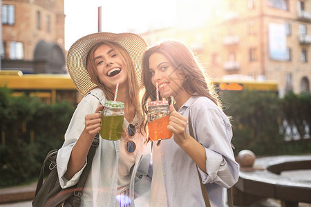 喝柠檬水两个穿着暑期服装的美丽微笑的年轻女子 在城里玩得很开心 喝着新鲜鸡尾酒 时髦 魅力背景