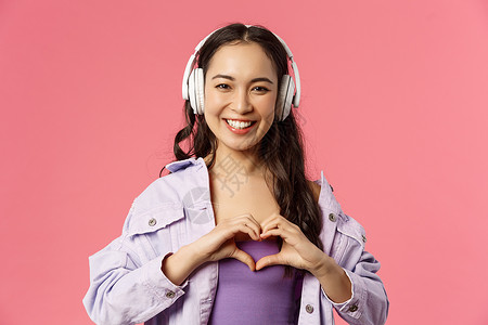 特写迷人 可爱的亚洲女孩戴着耳机听音乐 在胸前展示心形 爱上她最喜欢的艺术家 歌手表演很棒的新歌 粉红色背景 夏天 跳舞背景图片