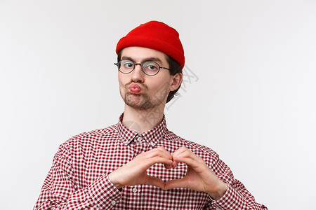 特写肖像滑稽的白人男性 长相古怪 戴眼镜和红色帽子 噘嘴要亲吻 表现出表白爱的心形 以卑鄙的方式表达同情 白色背景程序员高清图片素材
