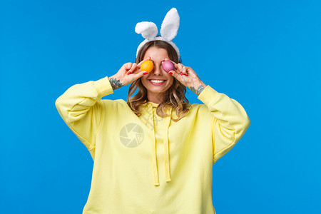假期 传统和庆祝概念 快乐无忧无虑地微笑着庆祝复活节的白人女性 眼睛上拿着两个彩蛋 咧嘴笑着 戴着可爱的兔耳 蓝色背景 脸 美丽女孩高清图片素材