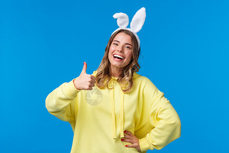 假期 传统和庆祝概念 乐观可爱的金发女孩 长着兔子耳朵 笑着摆出赞同的姿势 竖起大拇指称赞干得好 复活节快乐 蓝色背景美丽的高清图片素材